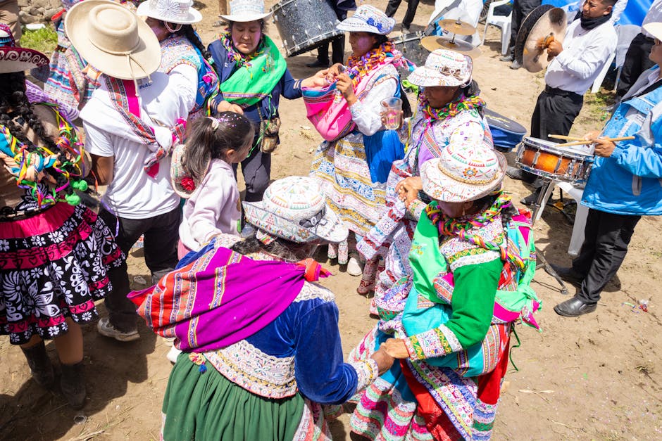 A group of people dressed in colorful traditional clothing, including embroidered skirts, shawls, and wide-brimmed hats, gathered outdoors on a sandy surface. They are engaged in a dance or ceremonial activity, holding hands and forming a circle. Some individuals are wearing patterned fabrics and accessories, while others are observing or participating. A few men are visible in the background, one holding a drum and another wearing a white shirt and dark trousers. The scene is lively, with bright sunlight illuminating the scene, and there are plastic chairs and utensils indicating a communal event or celebration. This cultural gathering captures the vibrant attire and cooperative spirit of the participants, suitable for documentation of community events or traditional festivities.
