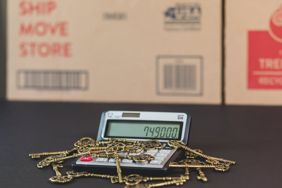 A calculator with a large digital display showing the number 749800 is positioned on a black surface, surrounded by several antique-style brass keys of various shapes and sizes, some attached to chains. In the background, there are several cardboard boxes with printed labels, including one that partially reads 'SHIP MOVE STORE' in red text and barcode markings. The scene appears to be set in a moving or packing environment, possibly inside a storage or loading area, with soft lighting highlighting the objects. The image illustrates aspects of home relocation, packing materials, and the logistical process of furniture transport, which aligns with the services offered by Man with Van Hayes in the removals category.