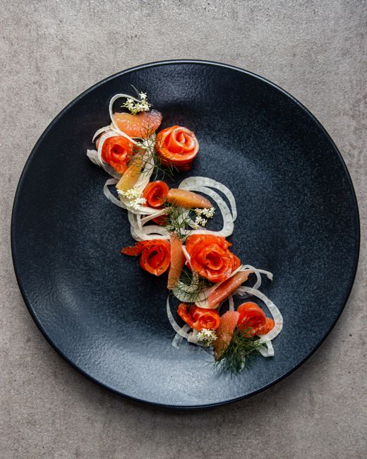 A black round plate on a grey surface holds a presentation of thinly sliced smoked salmon arranged in delicate rosettes, interspersed with finely sliced white onions and garnished with fresh dill spri