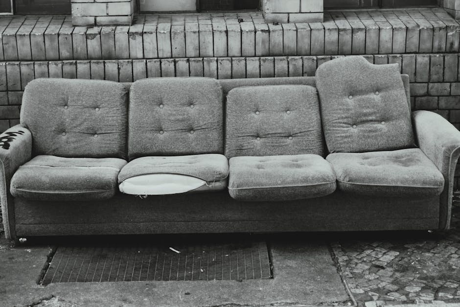 A black and white photograph showing a worn fabric three-seater sofa positioned outdoors on a pavement in front of a brick wall. The sofa has visible signs of use, with a torn and partially missing cushion on the left side, exposing the filling underneath. The middle cushion appears to have some creases, and the right armrest has noticeable wear. The sofa's fabric is textured, with button tufting on the backrest and seat cushions. To the left of the sofa, an armless section of the sofa is visible with a partially wrapped wheel or object. The sofa is situated on a concrete or stone surface, with a metal grate installed underneath part of it. This scene likely depicts a furniture collection effort or home relocation process coordinated by Man with Van Hayes, featuring furniture disassembly, packing, and transportation activities related to moving or furniture transport services.