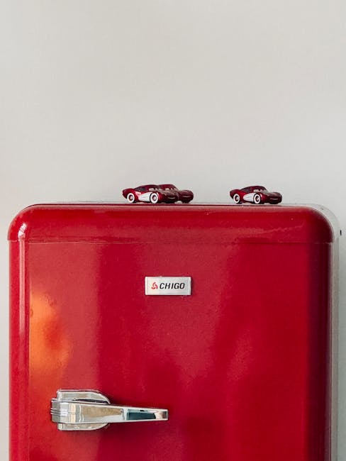 A red Chigo mini fridge with a silver handle is positioned indoors against a plain white wall, with two small toy race cars placed on top of it. The scene is well-lit, and the fridge appears to be in a residential setting, possibly in a room prepared for packing or moving. This setup reflects aspects of home relocation, with the fridge awaiting transport or part of a packing process. Man with Van Hayes specializes in removals, including the careful handling and transportation of household appliances and furniture, as seen here with the focus on the fridge and accompanying toys. The overall image captures a moment in the packing and moving sequence, emphasizing the importance of secure furniture and appliance transport within a household during house removals.