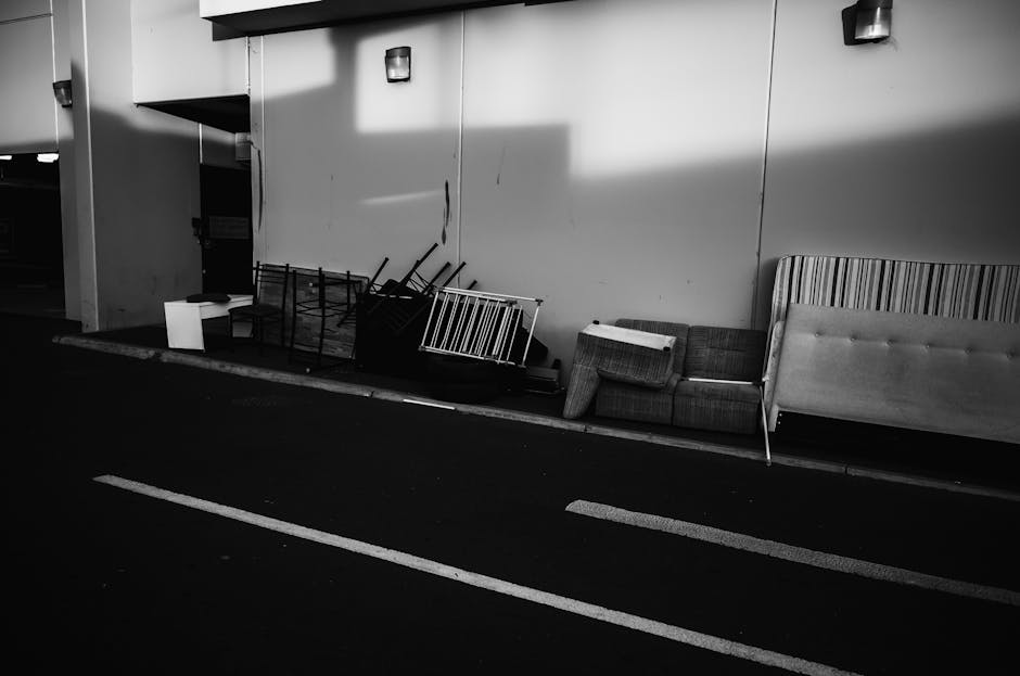 A black and white photograph showing a worn fabric three-seater sofa positioned outdoors on a pavement in front of a brick wall. The sofa has visible signs of use, with a torn and partially missing cushion on the left side, exposing the filling underneath. The middle cushion appears to have some creases, and the right armrest has noticeable wear. The sofa's fabric is textured, with button tufting on the backrest and seat cushions. To the left of the sofa, an armless section of the sofa is visible with a partially wrapped wheel or object. The sofa is situated on a concrete or stone surface, with a metal grate installed underneath part of it. This scene likely depicts a furniture collection effort or home relocation process coordinated by Man with Van Hayes, featuring furniture disassembly, packing, and transportation activities related to moving or furniture transport services.