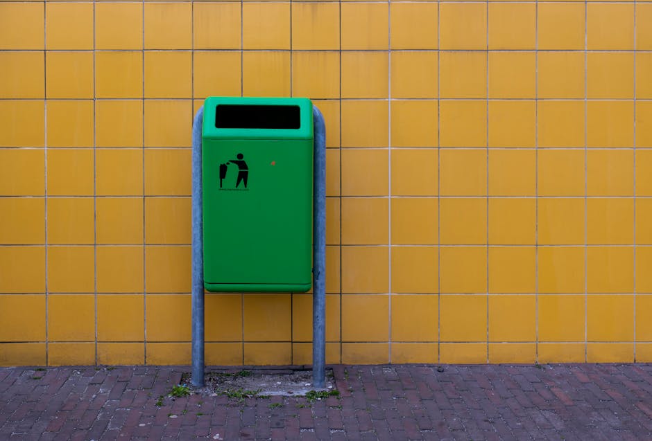 A green public waste bin with a black pictogram of a person disposing of rubbish is mounted on two grey metal poles, positioned against a yellow tiled wall outdoors. The bin is rectangular with a black opening at the top for waste disposal. The wall behind it features evenly spaced horizontal and vertical grout lines, creating a grid pattern, and the ground is paved with reddish-brown bricks. Small patches of grass and dirt are visible at the base of the poles. This setting illustrates the typical environment for waste disposal in a residential or commercial area, relevant to home relocation and packing processes managed by Man with Van Hayes, who provides removals and moving services, including waste disposal and recycling in accordance with local council regulations.
