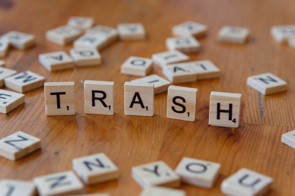 Photograph of scattered Scrabble tiles on a wooden floor, with the word 'TRASH' spelled out in uppercase letters using four tiles in the center. Around the word, additional tiles are randomly placed, some upside down or partially visible. The tiles are made of wood with black printed letters and small point values beneath each letter. The background includes a partially open doorway leading into an interior space, with a cardboard box and some packaging materials like bubble wrap and plastic wrap nearby, suggesting packing or decluttering activities related to house removals or home relocation. The overall scene highlights the process of clearing out unwanted items, with a focus on waste disposal and recycling as part of a home move managed by companies like Man with Van Hayes.