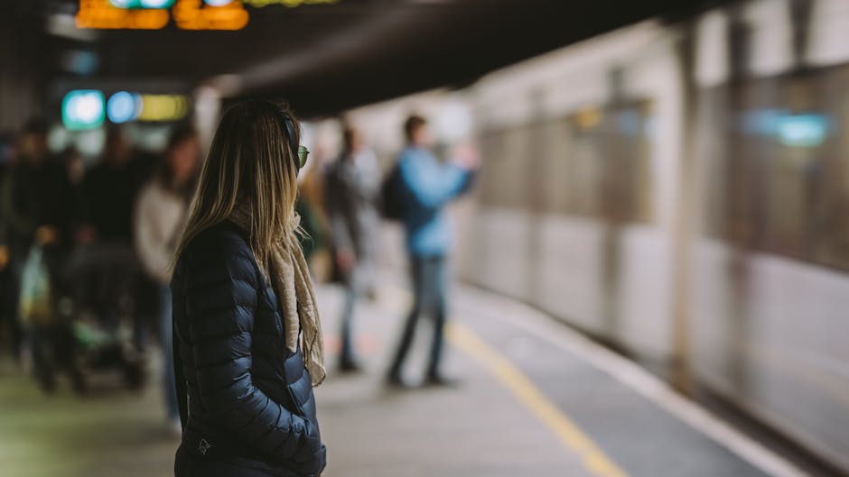 A woman with long blonde hair, wearing a dark blue puffer jacket and glasses, stands on a train station platform with her hands in her pockets, waiting for a train. In the background, several blurred passengers are seen, some standing and others walking along the platform, which has a yellow safety line along the edge. A silver train with closed doors is visible to the right, indicating a typical setting for transport or commuting. The station has an indoor environment with artificial lighting, and the scene appears to be during daytime. This image relates to house removals and relocation services, illustrating the transition and logistical considerations involved in moving home, such as packing, transporting furniture, and traveling between locations, as managed by professional services like Man with Van Hayes.