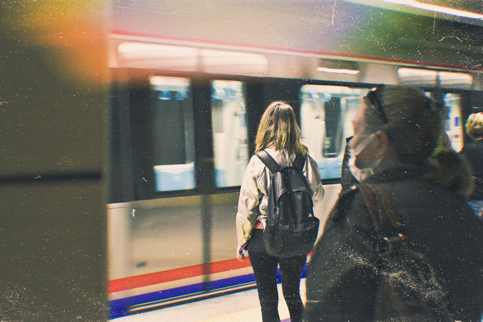 A woman with long blonde hair, wearing a dark blue puffer jacket and glasses, stands on a train station platform with her hands in her pockets, waiting for a train. In the background, several blurred passengers are seen, some standing and others walking along the platform, which has a yellow safety line along the edge. A silver train with closed doors is visible to the right, indicating a typical setting for transport or commuting. The station has an indoor environment with artificial lighting, and the scene appears to be during daytime. This image relates to house removals and relocation services, illustrating the transition and logistical considerations involved in moving home, such as packing, transporting furniture, and traveling between locations, as managed by professional services like Man with Van Hayes.