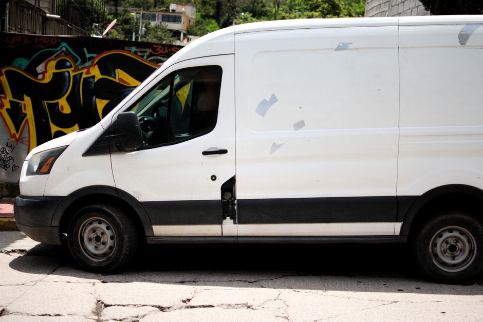 A white commercial van parked on the pavement outside a residential property, visible with its sliding side door slightly open, revealing a glimpse of interior packing materials and furniture items secured for moving. The van has black side mirrors and black protective trim along the lower edge, with a few minor scratches on the exterior. In the background, colorful graffiti art is painted on a wall, and a grassy area with trees and apartment buildings can be seen beyond. The scene captures the typical loading process involved in home relocation or furniture transport, with a focus on the vehicle used by Man with Van Hayes for house removals and moving services, alongside elements such as cardboard boxes, cushioning blankets, and protective wrapping materials visible inside and around the vehicle, indicating ongoing packing and transportation activities related to scheduled removals as per Hayes pavement and parking regulations.