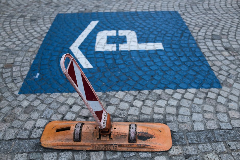 A close-up image of a designated parking space marked for disabled access on a cobblestone pavement, with a blue background and white wheelchair symbol painted on the ground. In the foreground, there is an orange and white striped barrier attached to a weighted base, positioned to block or protect the space. The barrier appears weathered, with visible black scuff marks and signs of rust. The ground around the space is composed of grey cobblestones arranged in a circular pattern. This scene is associated with loading or parking regulations relevant to house removals and furniture transport, possibly indicating restrictions or special access considerations. The presence of the barrier and markings suggests the area is prepared for safe vehicle loading, fitting within the context of a professional home relocation service provided by Man with Van Hayes, supporting efficient and compliant moving operations in Hayes.