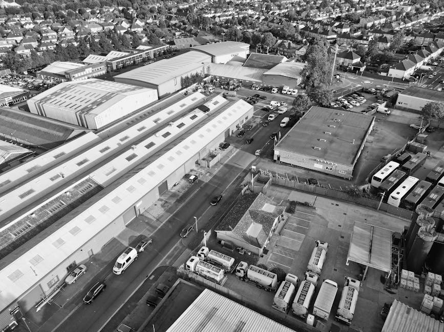 An aerial view of an industrial area and residential neighbourhood in Hillingdon, showing large warehouse-style buildings with flat and slightly pitched roofs, a parking lot filled with cars, vans, and trucks, some of which are likely used for home relocation and furniture transport services. In the foreground, there are smaller structures, including a small building with a pitched roof and adjacent parking spaces with several parked vehicles and vans. A wide road runs through the area, with cars and a white van marked with 'Man with Van Hayes' visible along the street, indicating active loading or unloading operations typical of moving logistics. The scene captures a mix of commercial and residential zones with houses and trees in the background, under daylight conditions with shadows illustrating the built environment and vehicles involved in packing and moving functions.