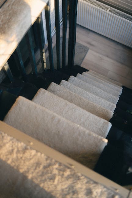 A view of a narrow staircase inside a residential building, with beige carpeted steps and a black metal railing on the left side. The staircase descends from an upper floor, which has wooden flooring partially visible at the top. To the right, a white radiator is mounted on the wall beneath a window, allowing natural light to illuminate the area. The image captures the confined space typical of flats in Hayes, highlighting the challenges of home relocation through narrow stairs. The scene is part of a moving process involving furniture transport, with no people or moving equipment visible, focusing solely on the staircase environment. This setting is relevant for house removals and packing and moving services, exemplifying the careful navigation necessary when carrying items through tight spaces, such as stairs in Hayes flats, in coordination with professionals like Man with Van Hayes.