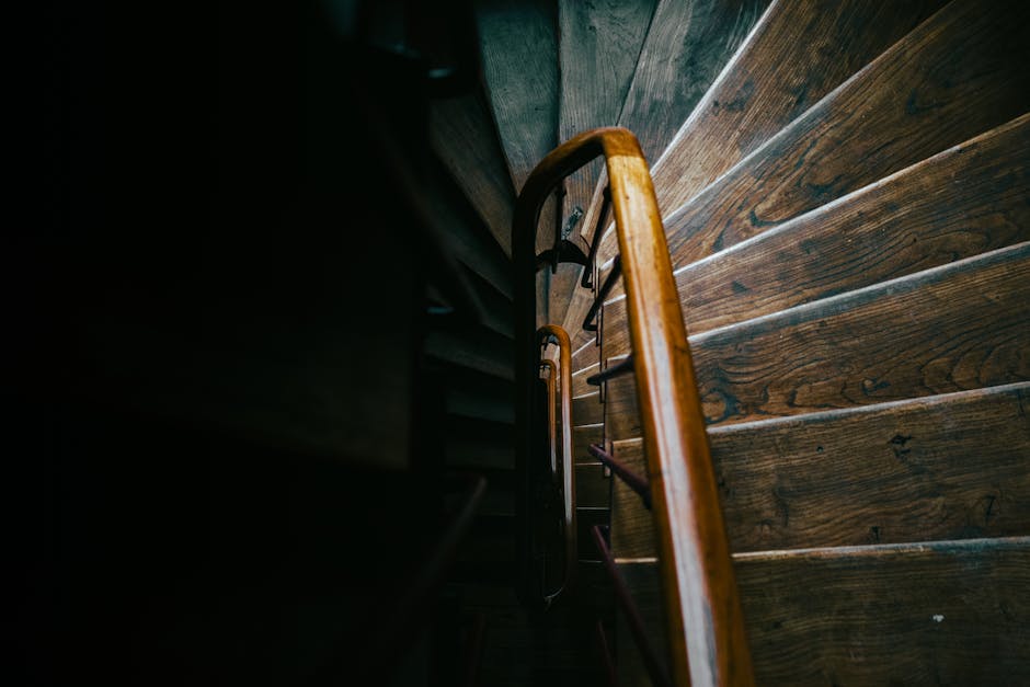 A downward view of a narrow staircase in a residential flat, with wooden steps and a polished wooden handrail on the right side. The staircase is enclosed by dark walls, with limited lighting creating shadows along the steps. The wooden treads exhibit a natural grain pattern, and the overall environment suggests an indoor setting within a house or apartment. The image captures the confined space typical of tight corridors or staircases in Hayes flats, which can present challenges for furniture transport and home relocation. As part of a moving process, the staircase may require careful planning for loading and unloading large items, such as boxes, small appliances, or furniture, potentially involving professional packing and moving services offered by Man with Van Hayes. The surrounding area is free of clutter, and no moving equipment is visible in this specific shot, focusing instead on the architecture of the staircase itself, essential for understanding the logistics involved in navigating limited space during a house move.
