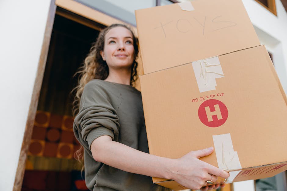 A woman with curly brown hair, dressed in a grey sweatshirt, is holding two large cardboard boxes, one stacked on top of the other, inside the doorway of a house. The boxes are taped and labeled, with the top one having the word 'toys' handwritten on it. She is standing on a paved area outside the property, which has a wooden frame around the entrance, and is looking slightly upward with a neutral expression. The interior of the house can be seen behind her, with storage shelves and various items. The scene depicts the process of home relocation, where she is engaged in packing or moving household belongings as part of a furniture transport or removal service by Man with Van Hayes, specialists in removals and emergency moving services for same-day relocations in Hayes.