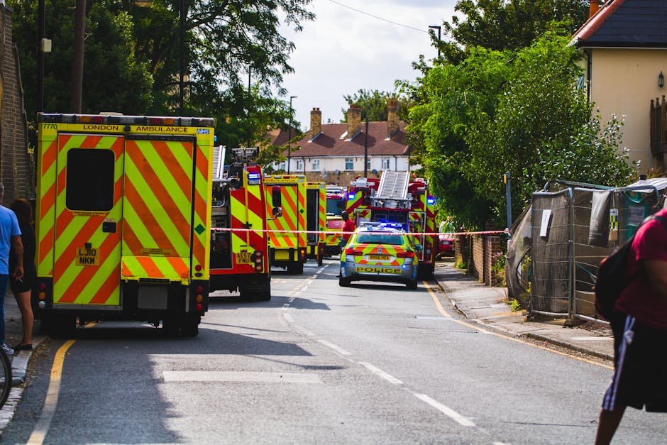 A scene on a residential street shows multiple emergency response vehicles, including a yellow and red London Ambulance and several fire engines, parked along the road with their lights flashing. The vehicles are positioned behind a cordon tape, restricting access to the area. In the foreground, there are a few pedestrians walking past, some partially visible on the sides of the image. The street is lined with trees, houses with brick and plaster facades, and construction fencing on one side. The emergency vehicles are loaded with equipment and are part of a coordinated response, likely involving planning and transportation of personnel and gear, typical of emergency services during a house evacuation or incident. The scene is well-lit with natural daylight, emphasizing the urban environment during a home relocation or incident response facilitated by [COMPANY_NAME], as part of the overall emergency or relocations process involving heavy vehicle transport and site safety measures.