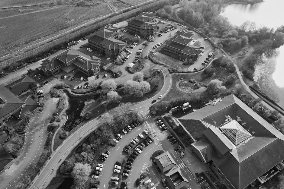 An aerial view in black and white shows a commercial office complex with multiple low-rise buildings featuring pitched roofs, surrounded by parking lots filled with numerous parked cars. Curved intern