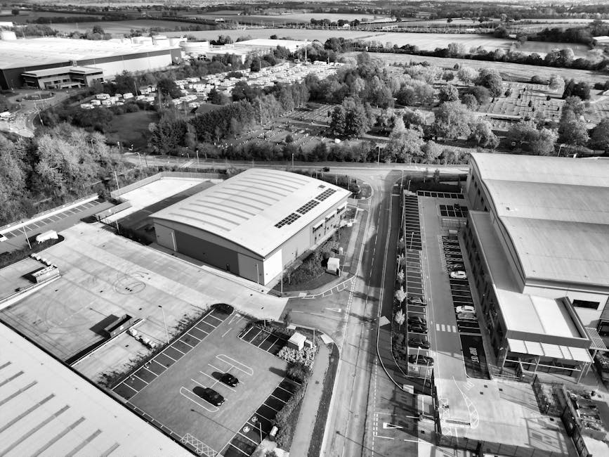 An aerial view in black and white shows a commercial office complex with multiple low-rise buildings featuring pitched roofs, surrounded by parking lots filled with numerous parked cars. Curved intern