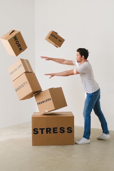A man dressed in a white T-shirt and blue jeans is depicted in a neutral indoor setting with plain white walls and a smooth concrete floor, engaged in a symbolic representation of stress related to house removals or moving logistics. He is standing next to a large cardboard box labeled 'STRESS' on the floor, with several other cardboard boxes around him, each labeled with words such as 'WORK', 'DEADLINE', 'ANXIETY', 'PROBLEMS', and 'BREAK UP', which are seen falling or being thrown into the air. The boxes are made of brown corrugated cardboard, with some covered in plastic wrap, and are positioned at various heights to illustrate the chaotic nature of packing and the emotional toll of home relocation. Visible on the left side of the image is an open flat surface, possibly a table, supporting some of the boxes. This scene visually captures the stress associated with moving, making it relevant for services like house removals and furniture transport offered by companies such as Man with Van Hayes, emphasizing the importance of professional packing and moving logistics to reduce stress during a home relocation process or furniture transport.