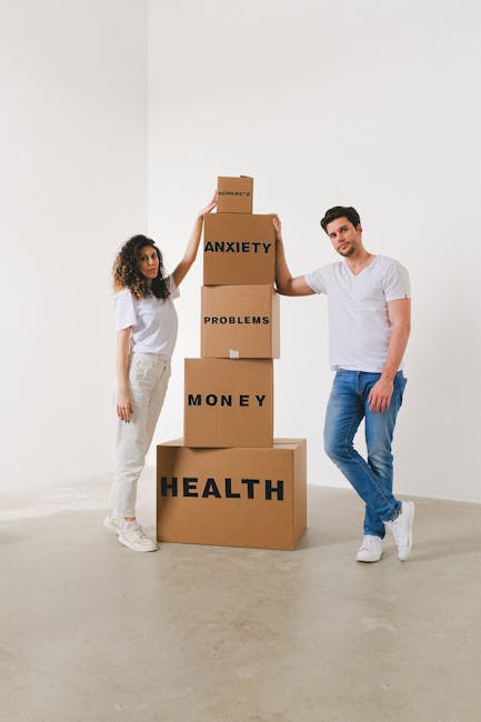 A man and a woman stand in a plain, well-lit indoor space with white walls and a light-colored floor. They are positioned next to a vertical stack of five brown cardboard boxes, each labeled with different words in bold black letters: the top box reads 'ANXIETY', the second box reads 'PROBLEMS', the third box reads 'MONEY', the fourth box reads 'HEALTH', and the bottom box reads 'HEALTH'. The woman, wearing a white t-shirt and beige pants, is standing to the left, gently holding the top box with her right hand, appearing to support or balance it. The man, dressed in a white t-shirt and blue jeans, is standing to the right with his right hand on his hip, and his left hand touching the stack of boxes, suggesting involvement in a moving or packing-related activity. The scene reflects an abstract representation of managing personal challenges, aligning with themes of packing and moving in the context of house removals as offered by Man with Van Hayes, emphasizing strength and support during home relocation or furniture transport.