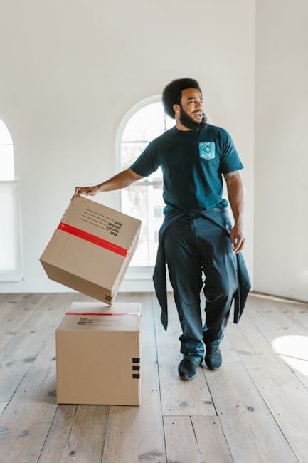 A man with dark curly hair and a beard, dressed in a navy blue t-shirt and dark trousers, is in a bright, empty room with white walls and wooden flooring. He is holding a cardboard box with a red tape seal, preparing for a house move, while balancing another box on top of a similarly packed box on the floor. The room features a large arched window allowing natural light to illuminate the space and the moving process. The man appears to be engaged in packing and furniture transport activities as part of a home relocation. Occasionally, a visible part of a moving company, Man with Van Hayes, may be implied through context, emphasizing professional removals and packing services. The scene illustrates the initial stages of loading boxes for a professional move, highlighting careful handling and the organization involved in packing and furniture transport during a flat-to-flat move, aligned with house removals processes.