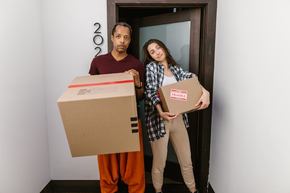 A man and a woman stand inside a doorway, each holding cardboard boxes for house relocation. The man, wearing a maroon t-shirt and orange trousers, holds a large box with a red and white shipping label. The woman, dressed in a checkered shirt and beige pants, carries a smaller box marked with a red 'Fragile' sticker. Behind them, the apartment door is partially open, revealing a narrow interior corridor with white walls. The scene is set in a well-lit indoor environment, indicating an ongoing furniture transport process involved in packing and moving activities, as managed by Man with Van Hayes. The boxes are made of brown cardboard, some with protective plastic wrap, and the individuals appear to be preparing for a flat-to-flat move, reflecting efficient loading and handling procedures typical of professional removals services.